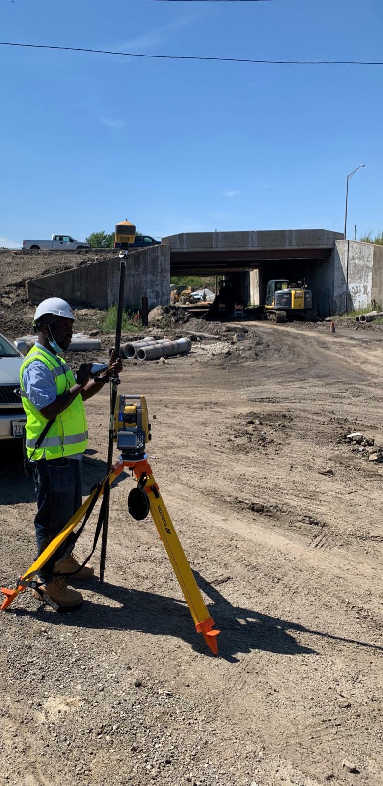 Engineer carrying out field surveying beside a total station and tripod near bridge construction works.